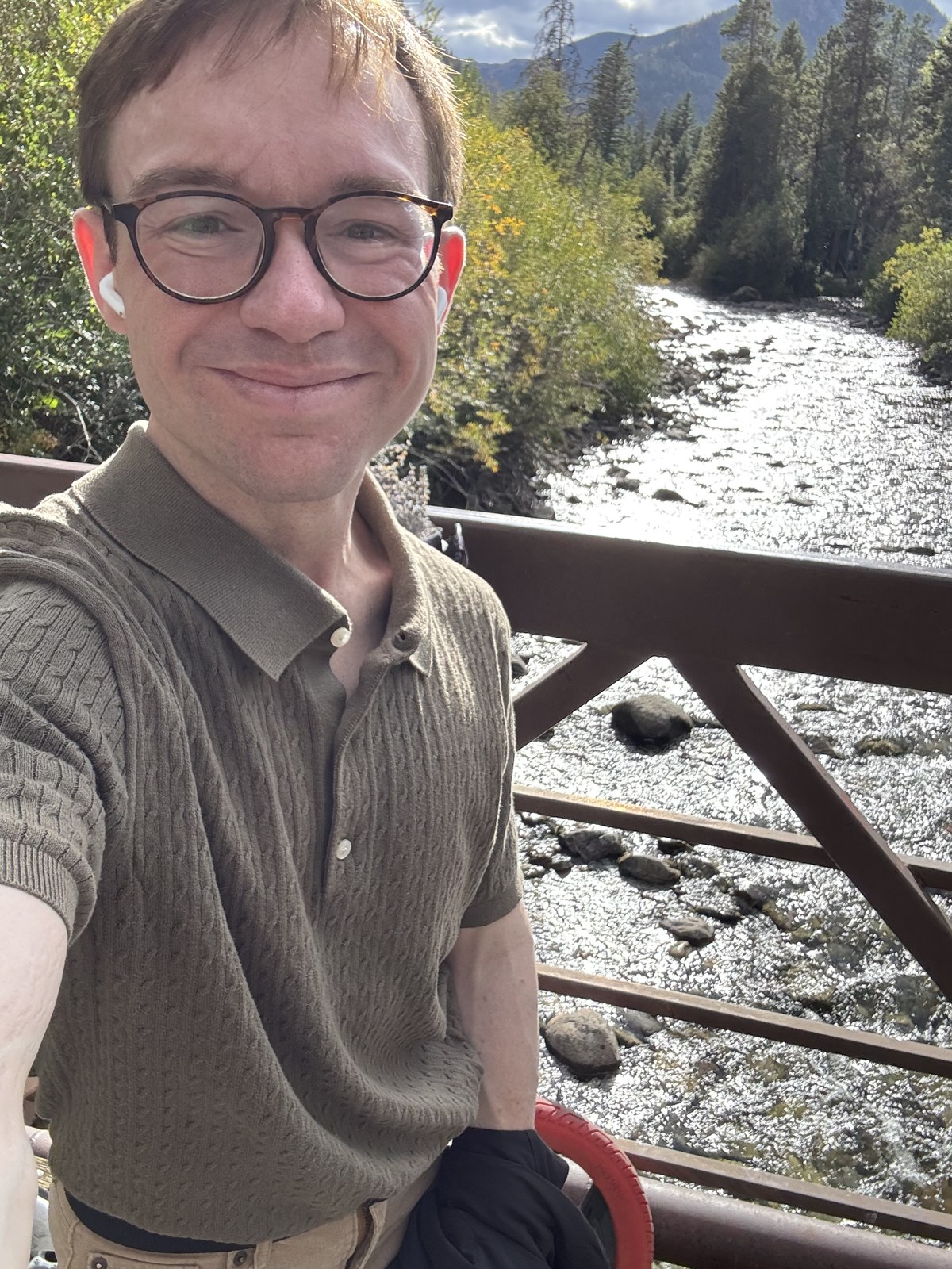 Avery Lane Maxwell, in glasses and an olive knit shirt, smiling on the Tenmile Creek footbridge with the creek and early-autumn trees behind him.